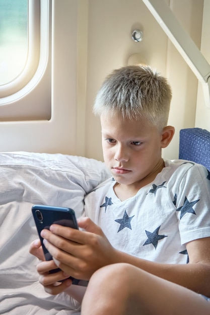 Teen boy reads electronic book with smartphone lying on white bedding on train car bottom shelf