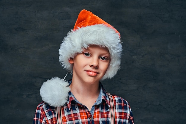 Teen boy in Santa's hat wearing a checkered shirt with suspenders. Isolated on a dark textured background.