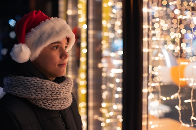 Teen boy in santas hat looking and dreaming in illuminated shop window xmas presents holidays or sho