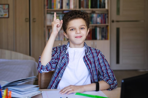 Teen boy shows his index finger up sitting at the table Training at home on coronavirus quarantine Selective focus Blur background