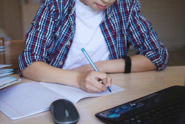 Teen boy at the table with a laptop thought and writes with a pen Coronavirus quarantined home schooling Selective focus Blur background