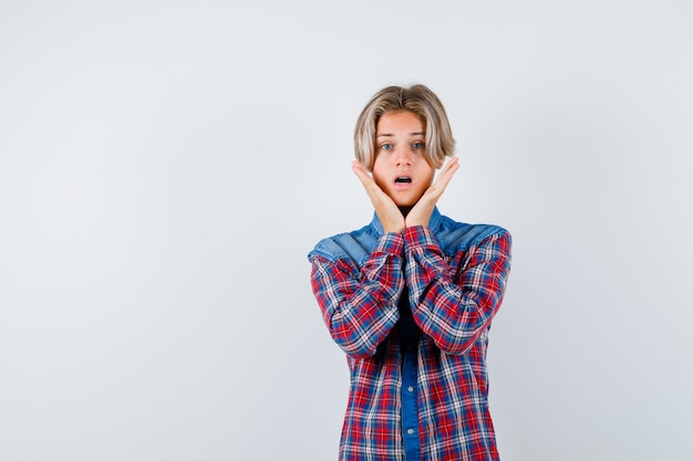 Teen boy with hands near cheeks in checkered shirt and looking perplexed. front view.