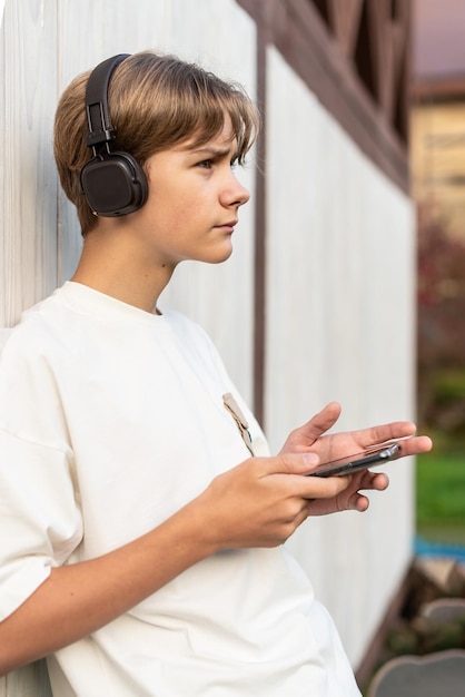 写真 スマートフォンで音楽を聴く10代の少年木製の白い背景で屋外で音楽を聞いています子供はスマートフォーンで楽しい時間を過ごし音楽を演奏しています テクノロジーのライフスタイルと人々のコンセプト