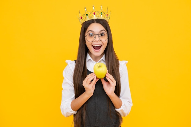 Teen child in queen crown hold apple isolated on yellow background princess girl in tiara teenage girl wear diadem excited face cheerful emotions of teenager girl