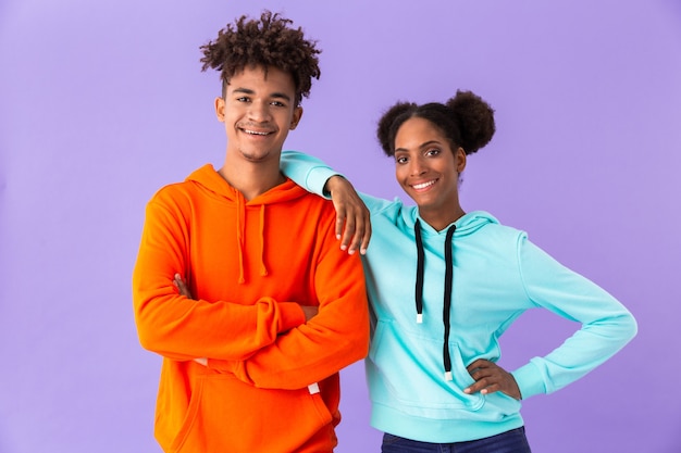 teen couple in colorful clothes smiling while standing together, isolated over violet wall