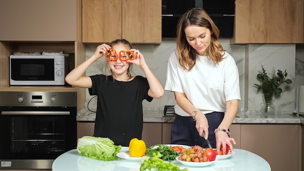 Teen daughter watches mother cutting red bell pepper on table with fresh vegetables and steals slices to imitate glasses in kitchen at home