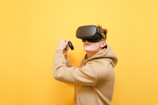 Teen gamer in a VR helmet stands on a yellow background with controls in his hands and plays virtual reality games looking away with a focused face Guy playing VR games with glasses isolated