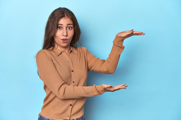 Teen girl in brown polo shirt on blue shocked and amazed holding a copy space between hands