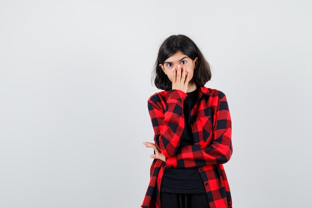 Teen girl in casual shirt covering mouth with hands and looking shocked , front view.