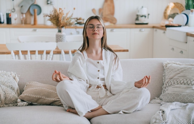 Teen girl in casual sitting in couch in lotus pose practicing meditation eyes closed at home