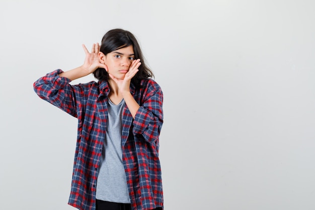 Teen girl in checkered shirt holding palms near mouth and behind ears and looking sensible , front view.