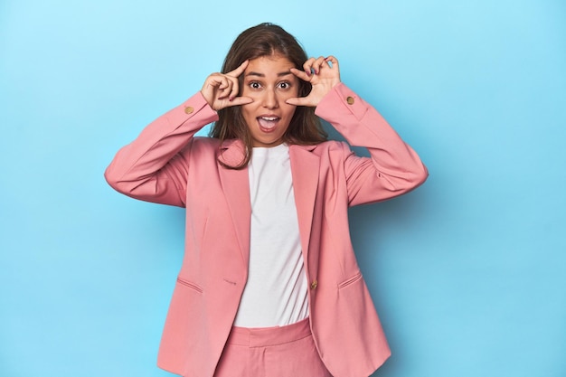 Teen girl in chic pink suit on a blue background keeping eyes opened to find a success opportunity