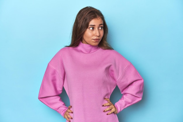 Teen girl in cozy pink sweatshirt on blue confused feels doubtful and unsure