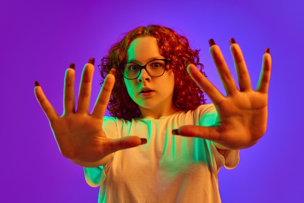 Photo teen girl emotional spreading hands with terrified face against purple background in neon light