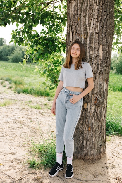 灰色のTシャツと木の近くの灰色のジーンズの十代の少女夏休みの自然散歩木に寄りかかる