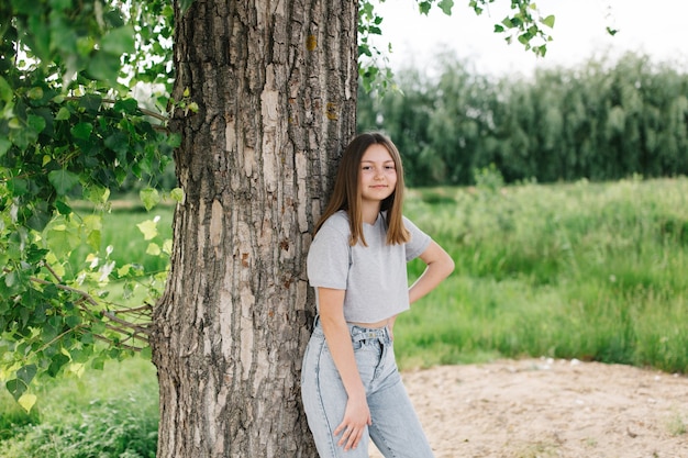 灰色のTシャツと木の近くの灰色のジーンズの十代の少女夏休みの自然散歩木に寄りかかる