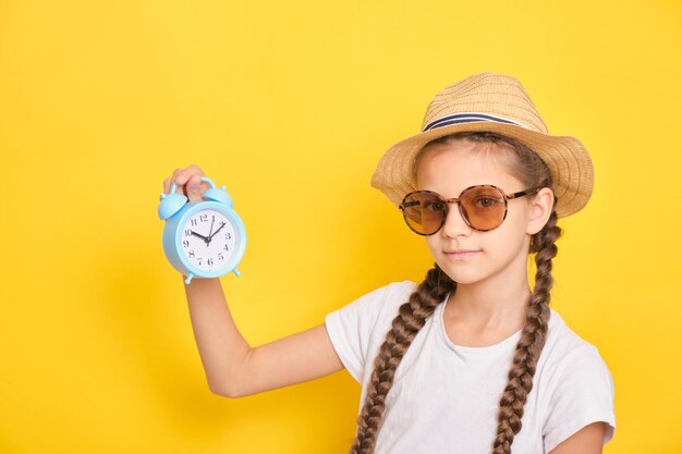 Teen girl in hat and sunglasses holds alarm clock on yellow background time to travel and vacation concept