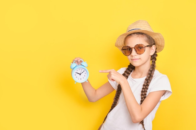 Teen girl in hat and sunglasses holds alarm clock on yellow background time to travel and vacation concept