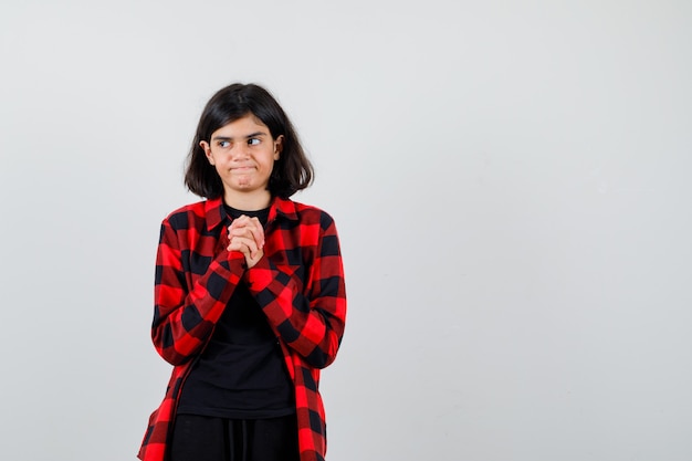 Teen girl holding clasped fingers over chest in t-shirt, checkered shirt and looking dissatisfied , front view.