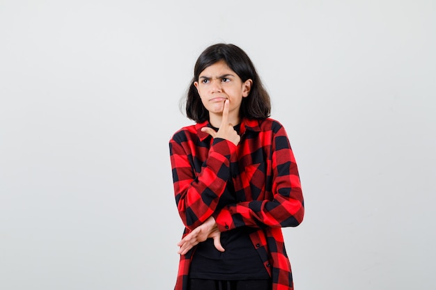 Teen girl holding finger near mouth in t-shirt, checkered shirt and looking thoughtful. front view.