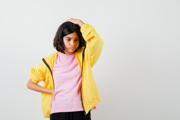 Teen girl holding hand on head and waist in yellow tracksuit, t-shirt and looking puzzled , front view.