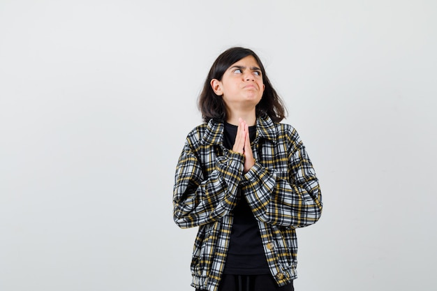Teen girl holding hands in praying gesture, looking aside in casual shirt and looking upset. front view.