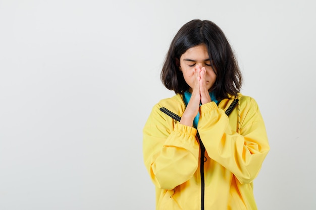 Teen girl holding hands in praying gesture in yellow jacket and looking hopeful , front view.