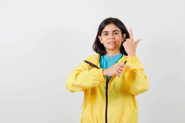 노란색 재킷을 입은 10대 소녀가 손가락을 위아래로 가리키고 유쾌한 모습을 하고 있습니다.
