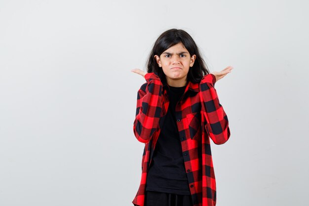 Teen girl keeping hands raised, shrugging shoulders in t-shirt, checkered shirt and looking disappointed , front view.