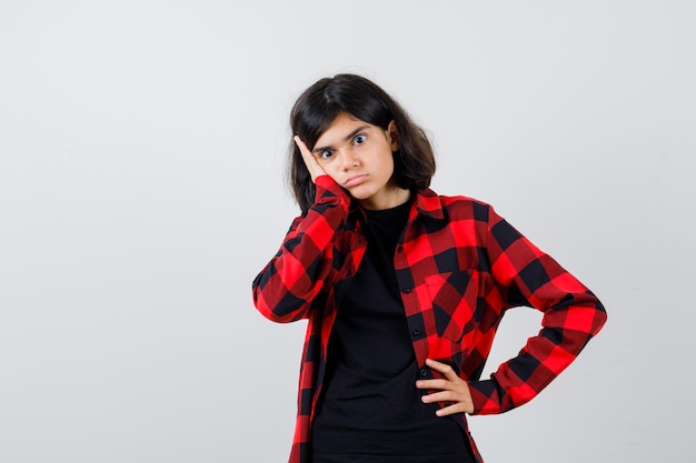 Teen girl leaning cheek on hand in t-shirt, checkered shirt and looking puzzled , front view.