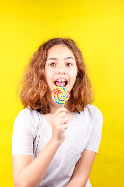 Photo teen girl licks a multi-colored round lollipop on yellow background