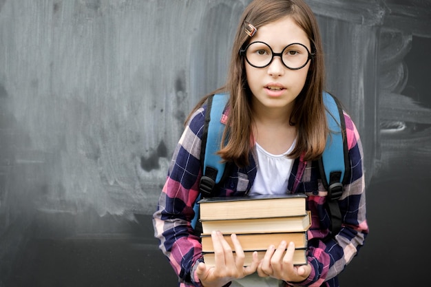 Teen girl little student holding pile of heavy books on
chalkboard background teenage girl in shirt glasses with blue
backpack and overweight stack of textbooks education school people
concept