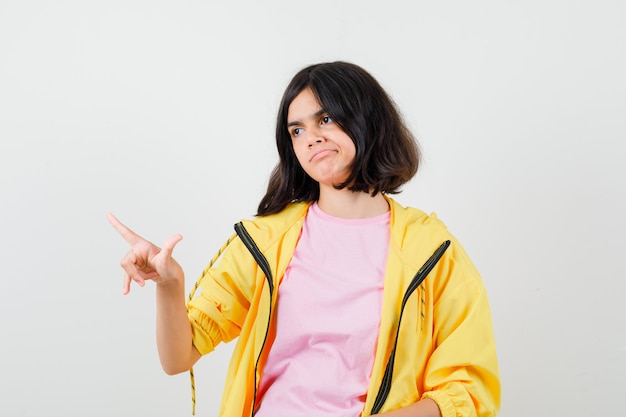 Teen girl pointing up, looking to the side in yellow tracksuit, t-shirt and looking upset, front view.