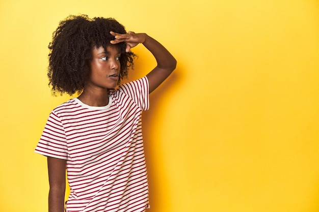 Teen girl in red striped Tshirt yellow studio backdrop looking far away keeping hand on forehead