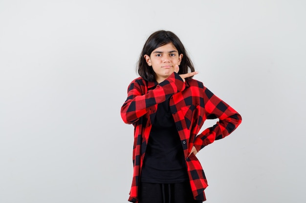 Ragazza teenager che mostra il gesto della pistola in camicia casual e che sembra perplessa, vista frontale.