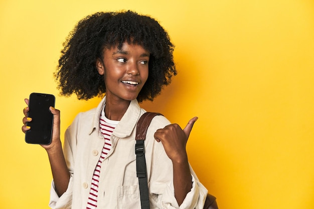 Teen girl showing smartphone backpack on yellow background points with thumb finger away