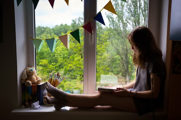 Ragazza teenager che si siede su un davanzale con un libro nella stanza dei bambini