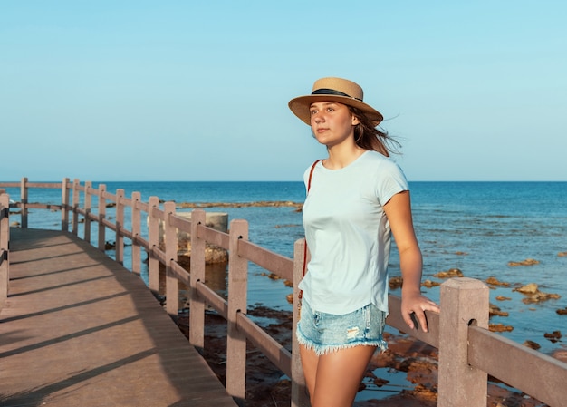 日没時に海沿いの木製の通路に立って、水色のTシャツ、麦わら帽子、財布を身に着けている10代の少女。夏の旅行のコンセプト