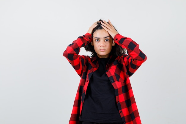 Ragazza teenager in t-shirt, camicia a scacchi che si tiene per mano sulla testa e sembra perplessa, vista frontale.