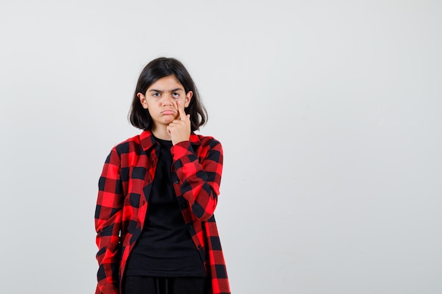 Teen girl in t-shirt, checkered shirt pulling down her eyelid and looking disappointed , front view.