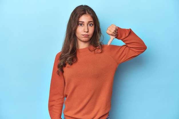 Teen girl in vibrant orange sweater on blue showing a dislike gesture thumbs down