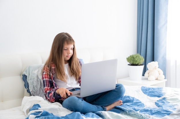 teen girl watching videos on laptop sitting on bed in her room. teenage school student studying online, distance learning,