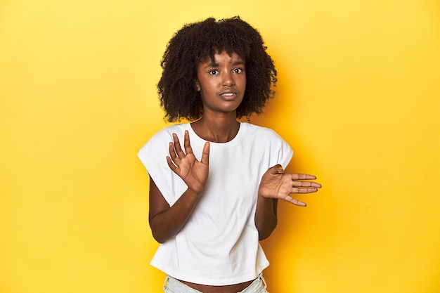 Teen girl in white tank top yellow studio background rejecting someone showing a gesture of disgust