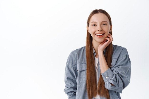 Teen girl with clean perfect skin touching cheek and smiling happily, got rid of acne, post cosmetics skincare treatment concept, standing pleased against white wall.