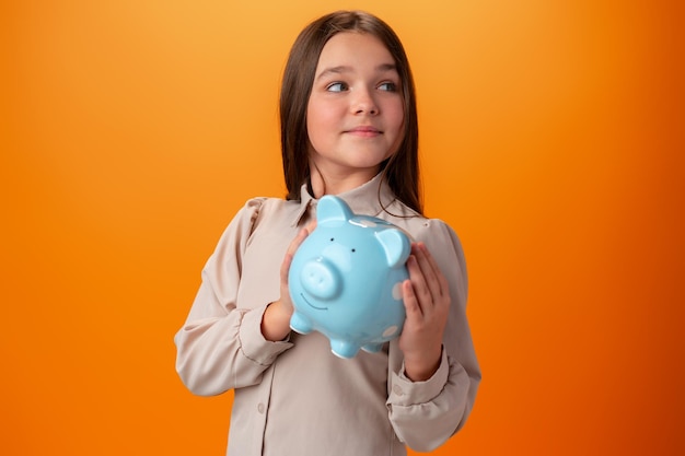 Teen girl with piggy bank on orange color background