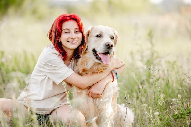 Teen girl with red hair hugging golden retriever dog at the nature and smiling Pretty young female with doggy pet in the sunny field