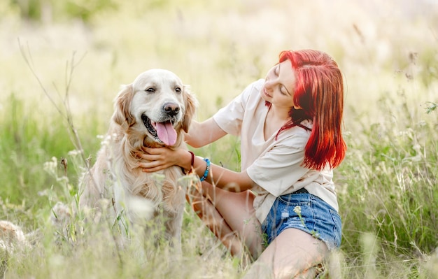 Teen girl with red hair hugging golden retriever dog sitting on the ground at the nature Pretty young female petting doggy pet in the sunny field