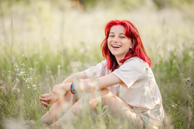 自然の中で地面に座って、手に花束を持って笑っている赤い髪の十代の少女夏の日当たりの良いフィールドでかなり若い女性