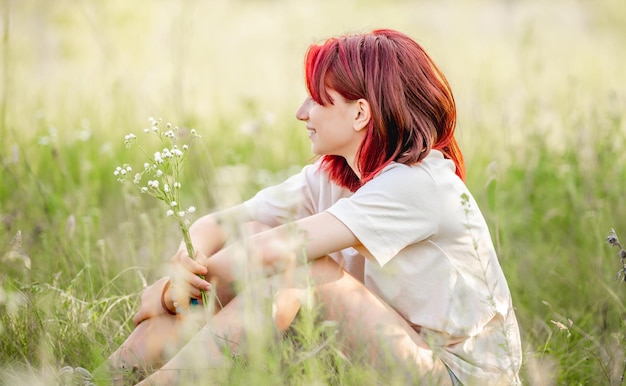 自然に座って、手に花束を持って笑っている赤い髪の十代の少女日当たりの良いフィールドでかなり若い女性