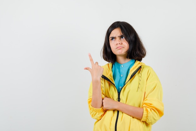 Teen girl in yellow jacket pointing up, looking away and looking thoughtful , front view.